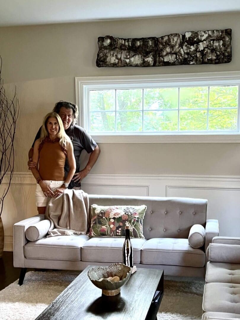 A couple standing by a window behind a beige couch in a cozy living room.