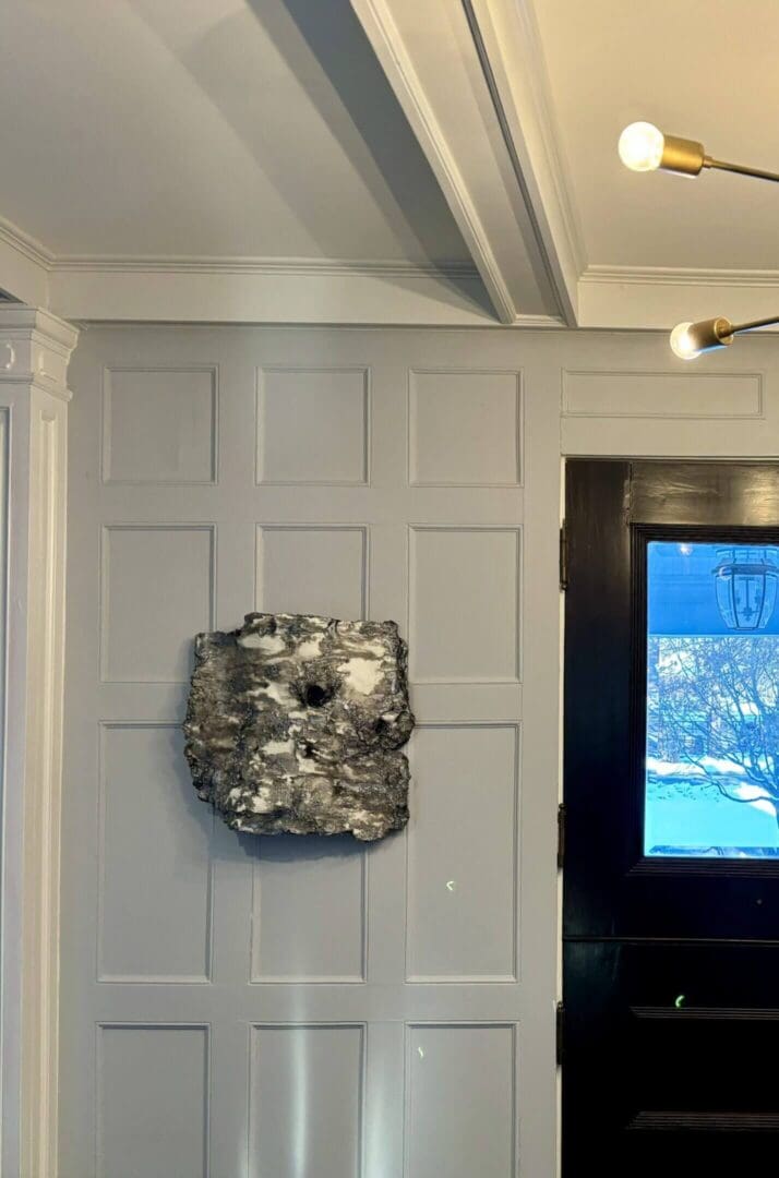 Modern art piece mounted on a paneled gray wall beside a window with a blue view.